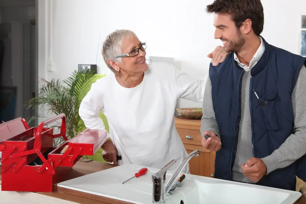 A handyman smiling while fixing a sink with an elderly lady standing next to him also smiling